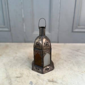 May include: A silver metal lantern with a handle. The lantern features a geometric design with cut-out patterns and a frosted glass panel. The lantern has a hexagonal base and an ornate design. The lantern is likely for decorative purposes.