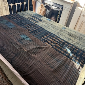 Può includere: Una trapunta patchwork con un design a righe e fantasia nei toni del marrone, blu e crema. La trapunta è stesa su una struttura letto in metallo bianco, mostrando la sua consistenza e le variazioni di colore. Il design della trapunta include toppe rettangolari.
