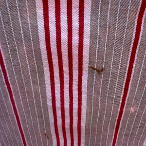 May include: Close-up of a fabric with a striped pattern. The design features alternating vertical stripes of red, white, and brown. The fabric has a textured appearance, with visible threads and slight imperfections.