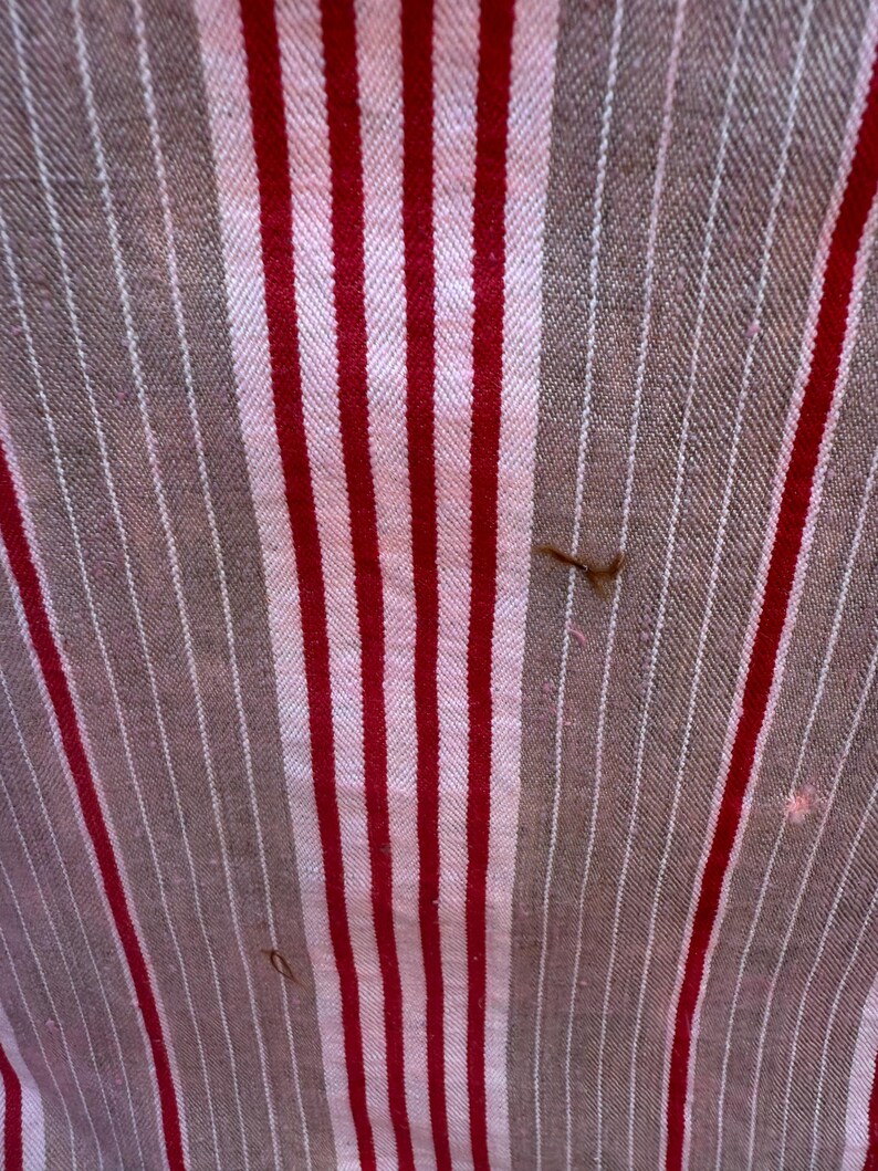 May include: Close-up of a fabric with a striped pattern. The design features alternating vertical stripes of red, white, and brown. The fabric has a textured appearance, with visible threads and slight imperfections.