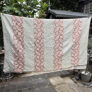 Puede incluir: Una colcha rectangular con paneles verticales alternos de tela color blanco roto y tela estampada con un diseño geométrico repetitivo en rojo terracota apagado. Toda la superficie muestra un acolchado denso y complejo.