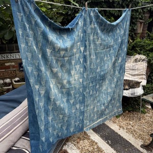 May include: A blue textile with a white geometric pattern of triangles and dots, hanging on a washing line. The fabric is draped, showing its texture and design. The background includes greenery and a brick wall.