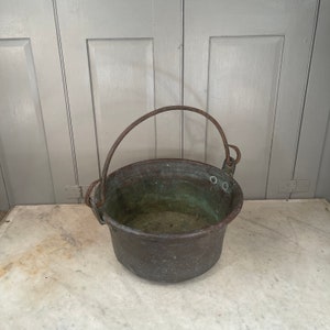 以下が含まれることがあります： 金属製の取っ手が付いたビンテージの銅製の鍋。鍋は緑青がかかっており、白い大理石の表面の上に置かれています。