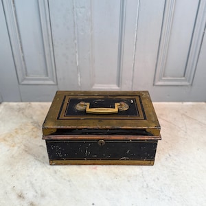 May include: A vintage black and gold metal box with a handle and lock. The box has a rectangular shape with a gold trim and a gold handle. The box is sitting on a white surface.
