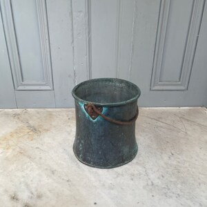 May include: A vintage copper bucket with a worn, green patina. The bucket has a metal handle with a heart-shaped detail.