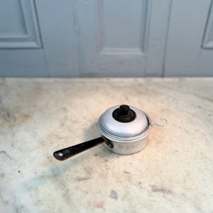 Op de afbeelding: Kleine, vintage aluminium steelpan met een zwarte handgreep en deksel. Het deksel heeft een zwarte knop. De steelpan staat op een marmeren oppervlak.