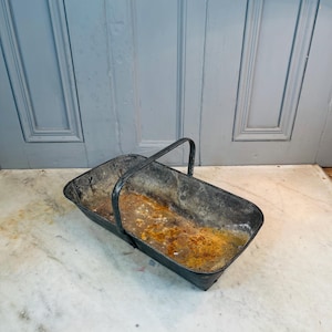 Puede incluir: Una cesta rectangular de metal desgastado con un solo asa. La cesta tiene un aspecto oscuro y envejecido con óxido y desgaste visibles. El asa es arqueada y está sujeta a los lados. La cesta está sobre una superficie de mármol.