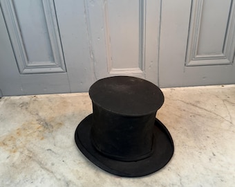 May include: A black top hat with a rounded crown and a wide brim. The hat is made of a dark material and sits on a marble surface. The hat is a classic formal accessory.