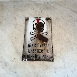 May include: A rectangular metal sign featuring a skull and crossbones symbol, and the Polish text "Nie dotykać! Urządzenie elektryczne!" in black. The sign has a distressed, aged appearance with rust and wear.