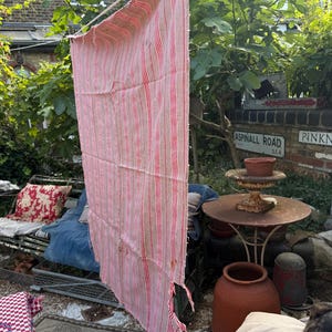 May include: A weathered, vertically-oriented textile with red and white stripes hangs outdoors. The fabric is frayed and shows signs of age. The textile is suspended from a line, suggesting it may be a curtain or a piece of vintage fabric.
