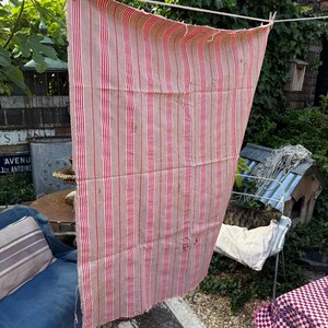 May include: A vintage, rectangular textile with vertical red stripes on a beige background, hanging on a clothesline. The fabric appears to be worn, with some visible imperfections. The textile is outdoors, with greenery in the background.