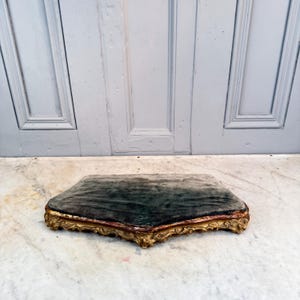 Puede incluir: Un taburete antiguo cubierto de terciopelo verde oscuro con un marco de madera dorado y ornamentado. El marco tiene una capa base roja. El taburete está colocado sobre una superficie de mármol. Medidas en centímetros.