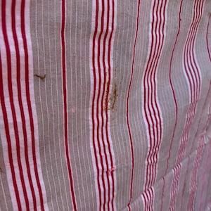 May include: Close-up of a fabric with vertical stripes in red, white, and gray. The fabric has a slightly wrinkled texture and some brown stains. The stripes are evenly spaced and create a classic pattern.