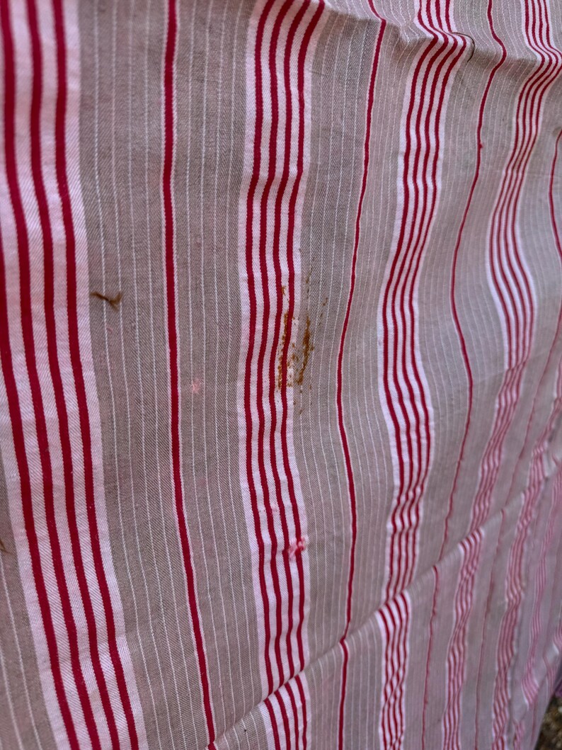 May include: Close-up of a fabric with vertical stripes in red, white, and gray. The fabric has a slightly wrinkled texture and some brown stains. The stripes are evenly spaced and create a classic pattern.