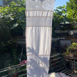 May include: A white crocheted tablecloth with a delicate floral pattern hangs on a clothesline in a garden setting. The tablecloth has a woven texture and is accented with beige stripes.