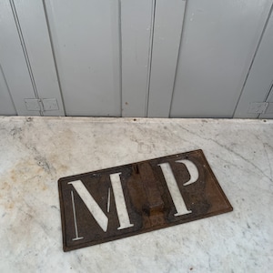 Può includere: Un cartello in metallo arrugginito con le lettere "M.P." tagliate in bianco. Il cartello è appoggiato su una superficie di marmo bianco.