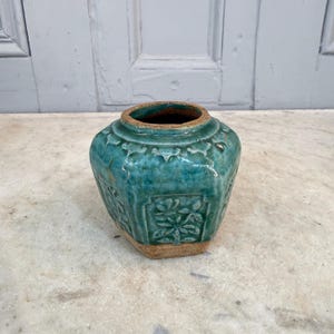 Pode incluir: Um jarro de cerâmica turquesa com base hexagonal e abertura larga. O jarro apresenta padrões florais em relevo e um acabamento envelhecido, dando-lhe um aspecto antigo. O jarro tem aproximadamente 15 cm de altura.