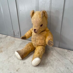 Puede incluir: Un oso de peluche vintage con pelaje marr&oacute;n claro, patas cosidas blancas y ojos bordados negros. El oso est&aacute; sentado sobre una superficie blanca.