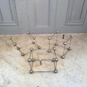 Puede incluir: Cuatro esculturas geométricas de color plateado. Cada escultura está hecha de finas varillas y esferas metálicas. Las formas incluyen cubos y formas abstractas. Las esculturas están dispuestas sobre una superficie de mármol.