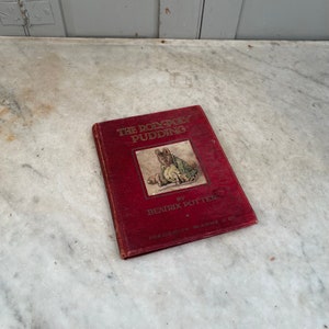 Pode incluir: Um livro de capa dura vermelho intitulado "The Roly-Poly Pudding" de Beatrix Potter. A capa apresenta uma ilustração de um rato vestindo uma jaqueta verde e segurando um rolo.