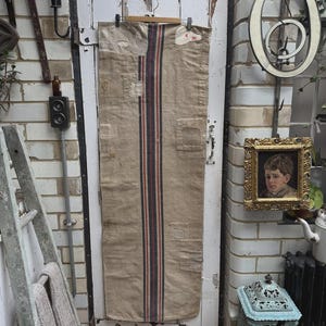 Può includere: Un sacco di grano in lino beige con una striscia verticale centrale rossa e blu navy. Appeso a una porta bianca, mostra una texture usurata e toppe, indicative della sua età e del suo utilizzo.