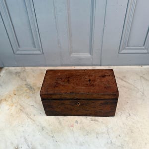 May include: A rectangular wooden box with a dark brown finish. The box has a small keyhole and appears to be antique. The box is sitting on a marble surface in front of a light blue door.