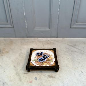 Puede incluir: Posavasos cuadrado antiguo con diseño floral. La baldosa central presenta flores azules y hojas verdes sobre un fondo crema, enmarcado por un borde dorado y un marco de metal oscuro. El posavasos tiene pequeños pies.