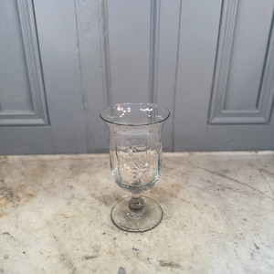 Op de afbeelding: Een helder glas met steel, een uitlopende bovenkant en een geëtst sterrenpatroon. Het glas staat op een ronde voet en heeft een delicate, vintage uitstraling. Het glas wordt waarschijnlijk gebruikt voor het serveren van dranken.