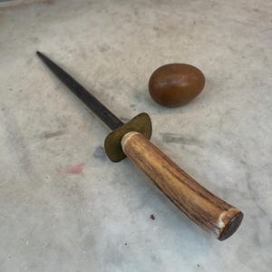Puede incluir: Un afilador de cuchillos de acero con mango de asta de ciervo marr&oacute;n y virola de lat&oacute;n. El afilador est&aacute; sobre una superficie gris claro junto a un objeto redondo marr&oacute;n.