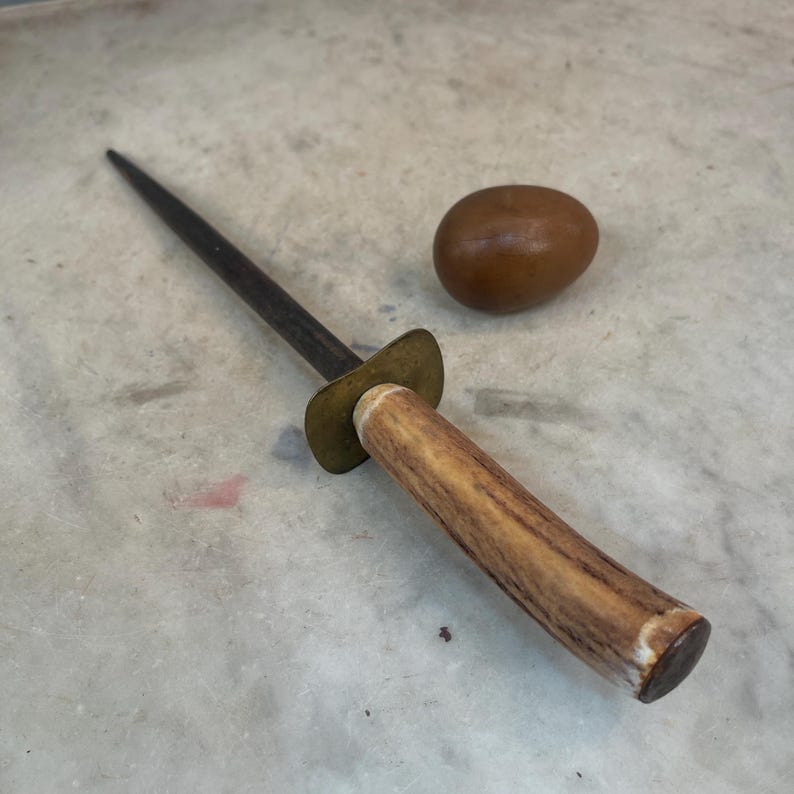 Puede incluir: Un afilador de cuchillos de acero con mango de asta de ciervo marr&oacute;n y virola de lat&oacute;n. El afilador est&aacute; sobre una superficie gris claro junto a un objeto redondo marr&oacute;n.