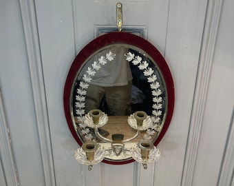 May include: Oval wall mirror with a burgundy velvet frame and etched leaf design. The mirror has four candle holders with crystal bases and brass accents. A decorative piece for home decor.