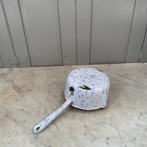May include: A white enamel saucepan with a blue and white speckled pattern and a red rim. The saucepan has a long, white handle.