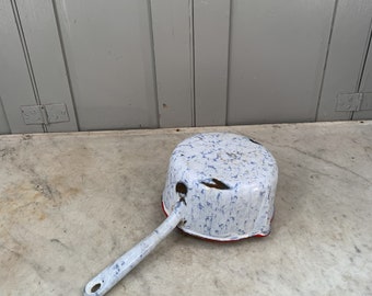 May include: A white enamel saucepan with a blue and white speckled pattern and a red rim. The saucepan has a long, white handle.