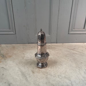 May include: A silver sugar shaker with a pierced top and a fluted base. The shaker is on a white marble surface.