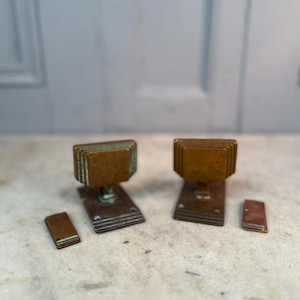 May include: Two antique bronze door knobs with rectangular, layered designs. Each knob is mounted on a rectangular base with a small, matching rectangular plate. The knobs and plates have a weathered, aged appearance.