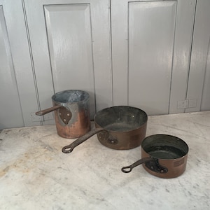 Puede incluir: Tres ollas de cobre con asas. Las ollas son de diferentes tamaños y tienen un aspecto desgastado y envejecido.