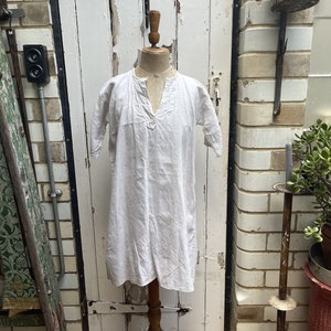 Può includere: Una tunica di lino bianco con scollo a V e maniche corte. La tunica è esposta su un manichino di legno contro un muro di mattoni bianchi.