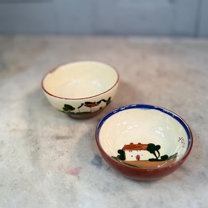May include: Two small, cream-coloured ceramic bowls with hand-painted designs. One bowl has a red rim and a landscape scene with a house and trees. The other bowl has a blue rim and a similar landscape scene with the word "you".
