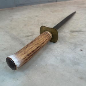 Puede incluir: Un afilador de cuchillos vintage de acero con un mango de asta de ciervo marr&oacute;n y un collar de lat&oacute;n. El afilador tiene una hoja larga y delgada.