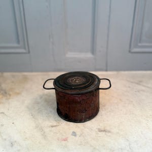 Puede incluir: Un pequeño recipiente cilíndrico antiguo con un exterior marrón rojizo oscuro y una tapa circular en capas. Dos asas pequeñas y curvas se extienden desde los lados. El recipiente está sobre una superficie clara.