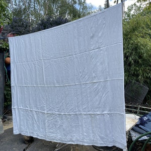 Può includere: Un tovaglia di lino bianco con dettagli ricamati, appesa a uno stendino all'aperto. La tovaglia presenta un motivo sottile di piccoli punti e un motivo ricamato più grande al centro.