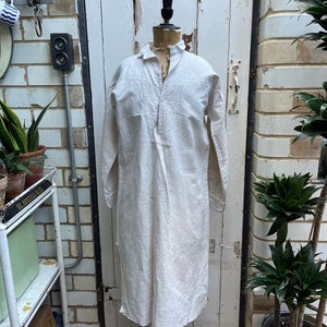 Peut inclure: Une chemise en lin blanc à manches longues et col boutonné. La chemise est sur un mannequin et est exposée contre un mur en briques blanches.