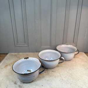 May include: Three white enamel bowls with black rims and handles. The bowls are slightly chipped and show signs of wear.