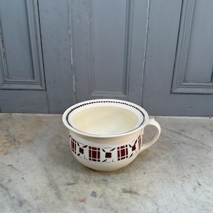 Puede incluir: Orinal de cerámica color crema con asa. El orinal presenta una banda decorativa de cuadrados rojos oscuros y diseños florales alrededor del exterior. El borde está acentuado con un patrón de puntos negros.