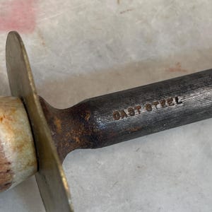 Puede incluir: Primer plano de un afilador de cuchillos de metal con mango de lat&oacute;n y las palabras "CAST STEEL" grabadas en el metal.