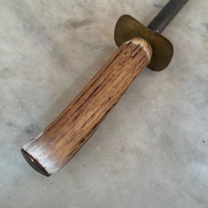 Puede incluir: Un afilador de cuchillos de acero vintage con mango de madera. El mango es de madera marr&oacute;n claro con una punta blanca. La parte met&aacute;lica del afilador es plateada y tiene una tapa de lat&oacute;n.