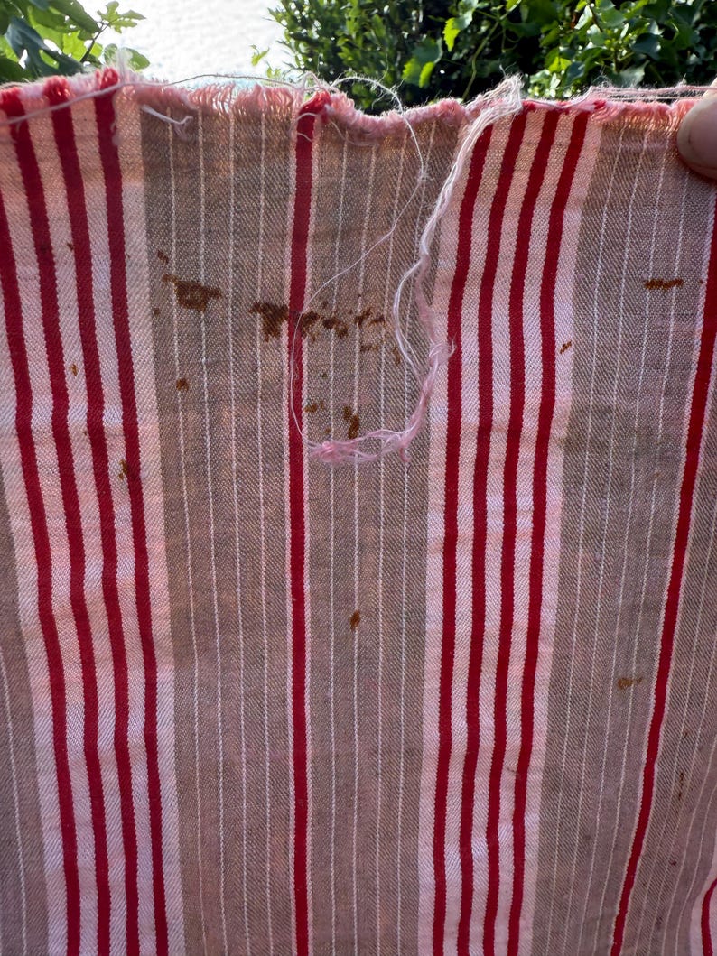 May include: Close-up of a striped fabric with red and white vertical stripes on a tan background. The fabric has a frayed edge and some brown stains. The fabric appears to be vintage or distressed.