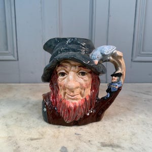 Puede incluir: Una taza de cerámica vintage con forma de cabeza de anciano, con barba roja y sombrero oscuro. Un pájaro azul se posa en el sombrero y una pequeña figura adorna el asa. La taza está pintada con rasgos faciales detallados.