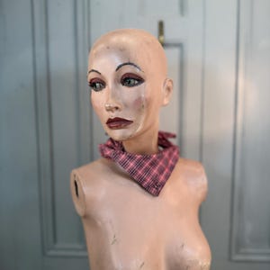 May include: A vintage mannequin head with painted facial features, wearing a pink and black plaid bandana. The mannequin has a distressed, aged appearance, with visible paint wear. The background is a light blue door.