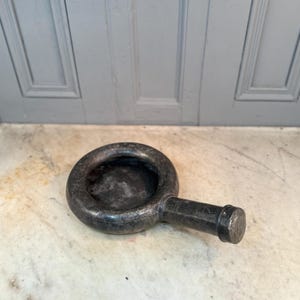 May include: A dark grey mortar and pestle. The mortar has a circular bowl and a handle. The pestle is cylindrical with a rounded end. The set is made of a dark, possibly stone, material.